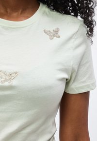 Κοντινό πλάνο ενός ατόμου που φοράει ανοιχτοπράσινο t-shirt με δύο διακοσμητικά πεταλούδες από στρας στην περιοχή του στήθους.