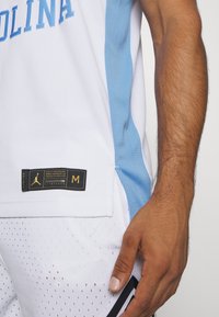 Maillot de basket-ball blanc avec des accents bleu clair, présentant une texture en maille, le texte "CAROLINA" et une étiquette noire avec un logo doré sur l'ourlet.