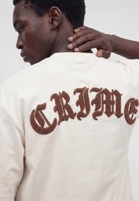 Homme à la peau foncée et aux cheveux courts et bouclés portant une chemise blanche avec de grandes lettres brunes texturées dans le dos indiquant "CRIME."
