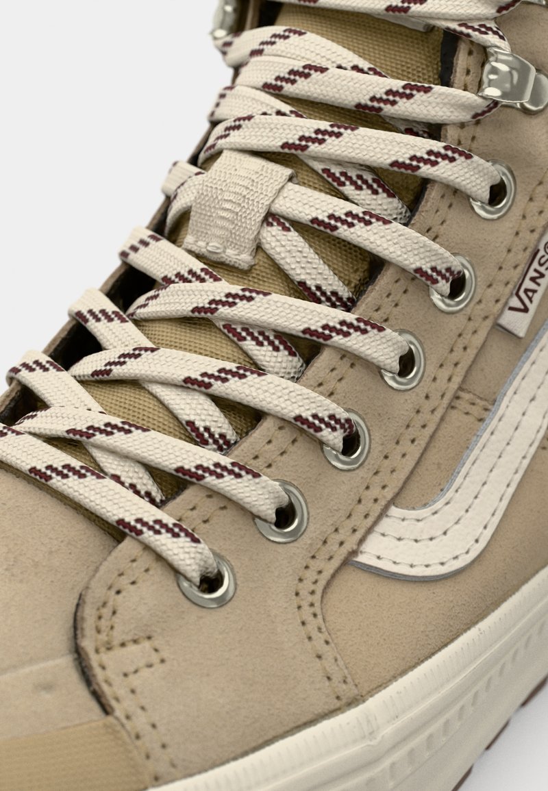 Zapatillas de ante beige con cordones blancos y burdeos, ojales plateados y una franja lateral blanca. Presenta una parte superior texturizada y suela de goma.