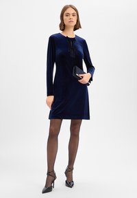 Marineblaues Samtkleid mit langen Ärmeln, rundem Ausschnitt und schwarzen Bandakzenten. Kombiniert mit transparenten Strumpfhosen und schwarzen High Heels, hält eine Clutch.