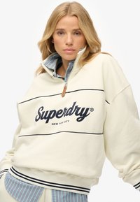 Cream-colored zip-up sweatshirt with navy stripes and "Superdry" logo, featuring a high collar and ribbed cuffs, layered over a plaid shirt.