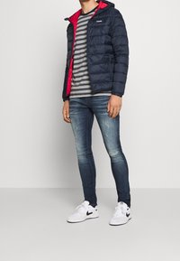 Navy puffer jacket with red inner lining, grey and white striped shirt, blue skinny jeans, and white sneakers with black accents.