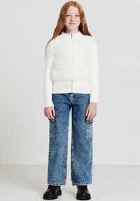 Giacca bianca con zip e un pannello morbido, abbinata a jeans a gamba larga di un blu chiaro con tasche frontali e un motivo a macchie. Scarpe platform nere.