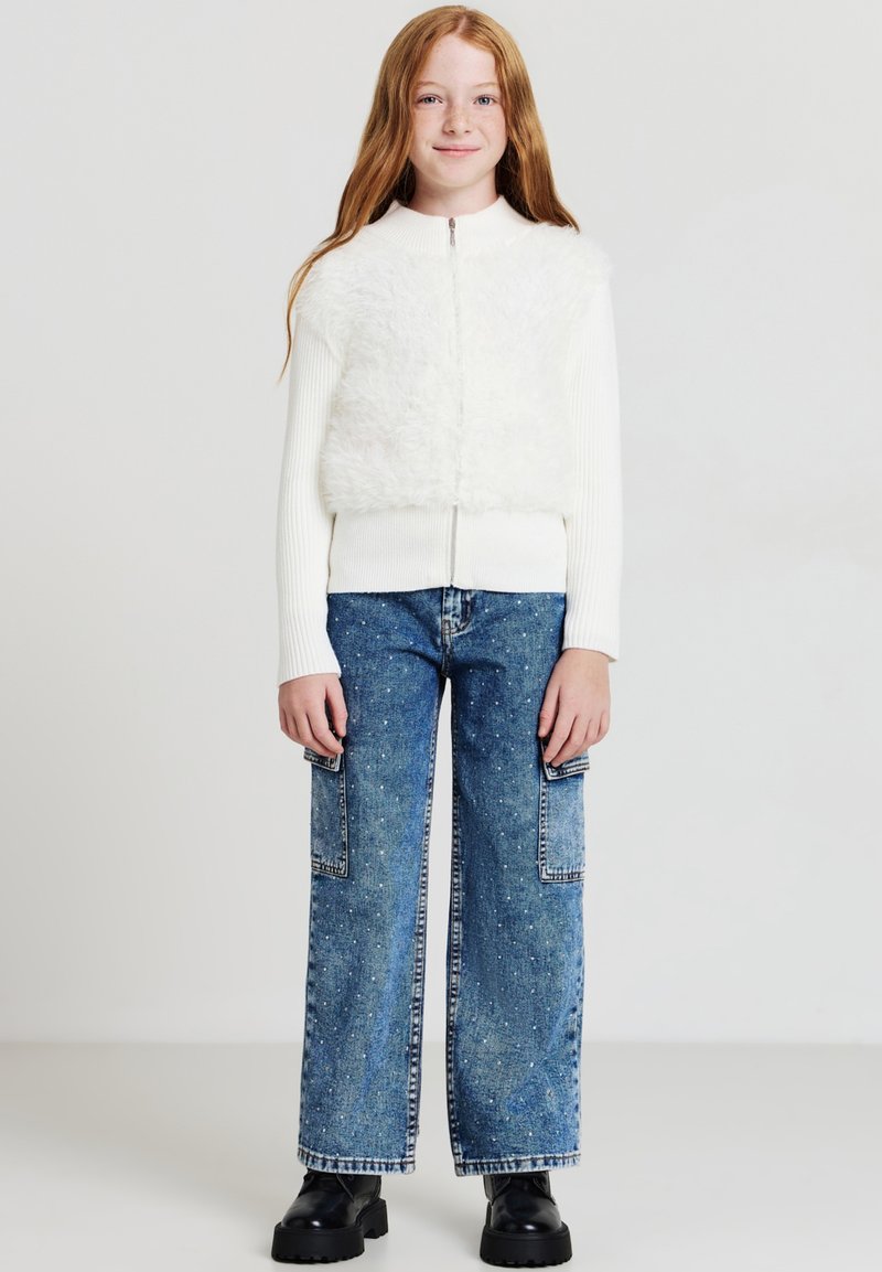 Giacca bianca con zip e un pannello morbido, abbinata a jeans a gamba larga di un blu chiaro con tasche frontali e un motivo a macchie. Scarpe platform nere.