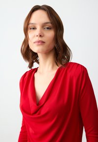 Czerwona bluzka z dekoltem w kształcie drapowania i długimi rękawami. Wykonana z gładkiego materiału o subtelnym połysku, z delikatnymi marszczeniami na ramionach.