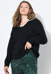 Jonge vrouw met golvend haar, gekleed in een zwarte geribbelde trui met witte rand en groene broek met paisleypatroon, poserend tegen een witte muur.