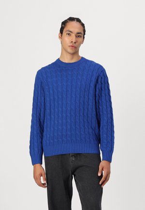 Pull bleu en tricot avec un motif de câble texturé, encolure ras du cou et ourlet côtelé. Porté avec un pantalon gris foncé, avec des manches longues.