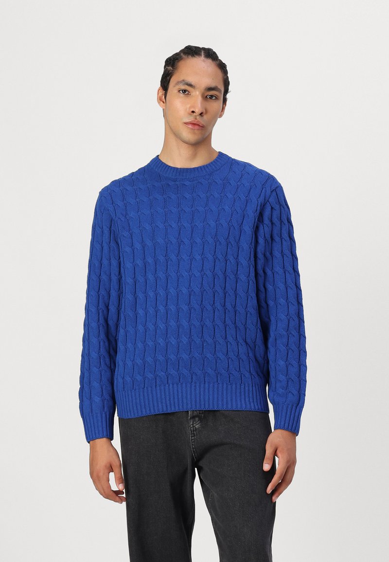 Blue knitted sweater with a textured cable pattern, crew neckline, and ribbed hem. Worn with dark gray pants, featuring long sleeves.