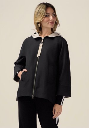 Kobieta w czarnej kurtce na suwak z beżowym kapturem i złotymi kolczykami kołami, patrzy w bok z jedną ręką w kieszeni.