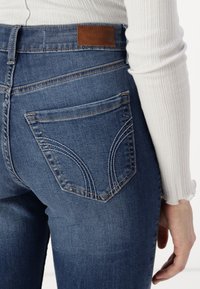 Σκούρα μπλε τζιν παντελόνι με στενή γραμμή, με καφέ δερμάτινη ετικέτα, δύο πίσω τσέπες και αντίθετες ραφές.