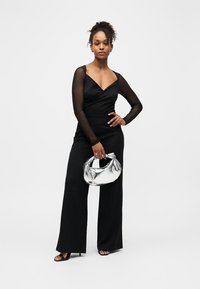 Femme portant une combinaison noire avec des manches longues en voile transparent, tenant un sac à main argenté, debout avec une main sur la hanche sur un fond uni.