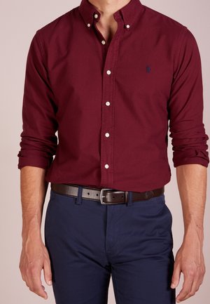 Homme portant une chemise bordeaux à manches longues boutonnée avec les manches retroussées, un pantalon bleu marine et une ceinture marron foncé.