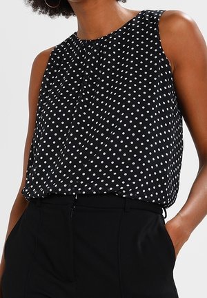 Donna che indossa una canotta nera senza maniche con pois bianchi infilata in pantaloni neri a vita alta, una mano in tasca.