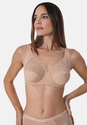 Frau trägt einen beigen, stützenden BH mit durchscheinenden, gemusterten oberen Körbchen und passende High-Waist-Slips, steht vor einem einfarbigen Hintergrund.