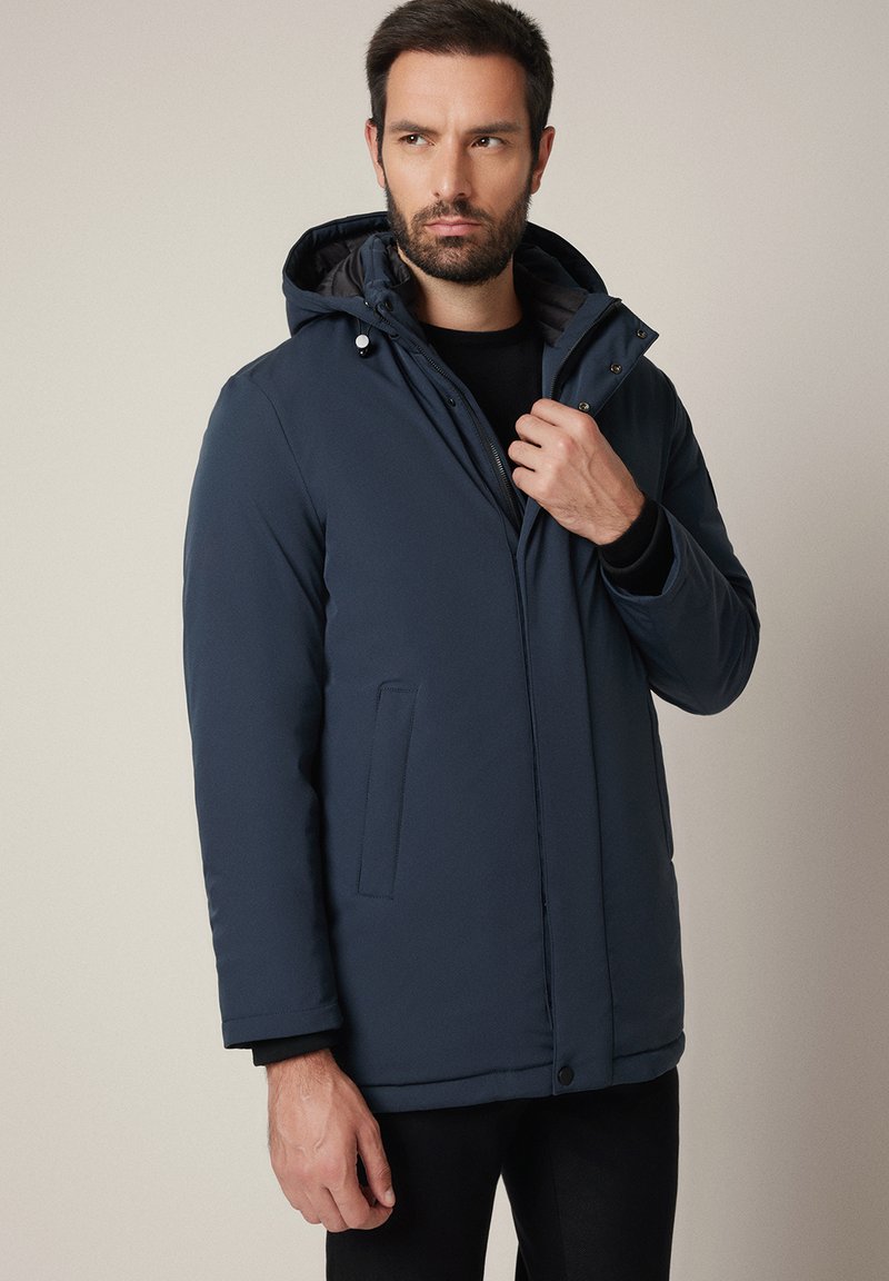 Mężczyzna w granatowej zimowej kurtce z kapturem, z zamkiem i napami, poprawiający kołnierz, stojący na jednolitym jasnym tle.