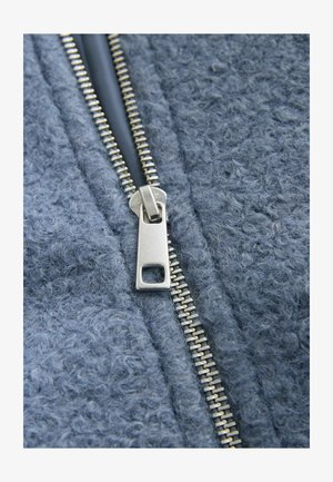 Šedá fleecová tkanina s textúrovaným povrchom, so strieborným kovovým zipsom uprostred na zapínanie.