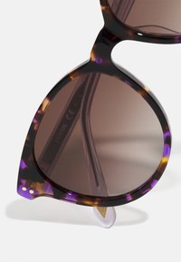 Sonnenbrille im Schildpattdesign mit einem markanten lila Muster, braunen Gläsern und einer auffälligen, übergroßen Cat-Eye-Form. Metallakzente im Gestell.