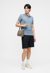 Άνδρας που φορά γαλάζιο πόλο μπλουζάκι, μαύρο σορτς, λευκές κάλτσες, κρεμ αθλητικά παπούτσια, κρατώντας μπεζ τσαντάκι χιαστί, στέκεται μπροστά σε λευκό φόντο.