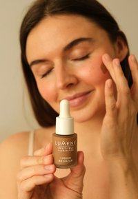 Bruine vloeibare bronzer in een glazen druppelfles met een witte druppelkap. Etiket leest "LUMENE INVISIBLE ILLUMINATION LIQUID BRONZER."