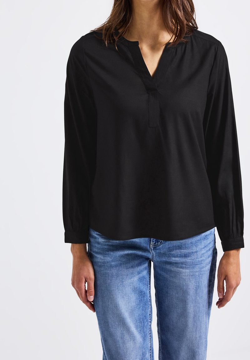 Femme portant un chemisier noir à manches longues avec un col en V et un jean bleu en denim, debout devant un fond clair uni.