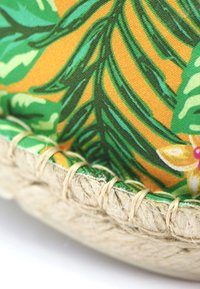 Coussin à motif floral avec des feuilles vertes et un fond jaune-orange. Bordé de fibre de jute naturelle. Surface de tissu texturée.