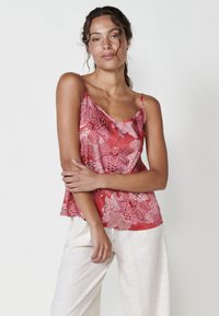 Camisole à motifs floraux rouges avec des accents en dentelle, en tissu léger, et bretelles spaghetti ajustables, assortie à un pantalon en lin de couleur crème.