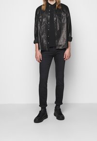 Chaqueta camisa de cuero negro con dos bolsillos frontales, combinada con un polo negro, jeans ajustados negros y zapatillas negras.