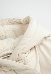 Puchowa beżowa kurtka z miękką, kremową podszewką z polaru i dużym kołnierzem. Posiada pikowaną zewnętrzną warstwę oraz subtelne detale przeszyć.