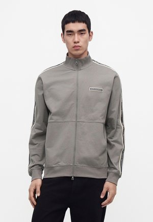Homme portant une veste grise zippée avec un col haut et des bandes de marque le long des manches, se tenant devant un fond blanc uni.