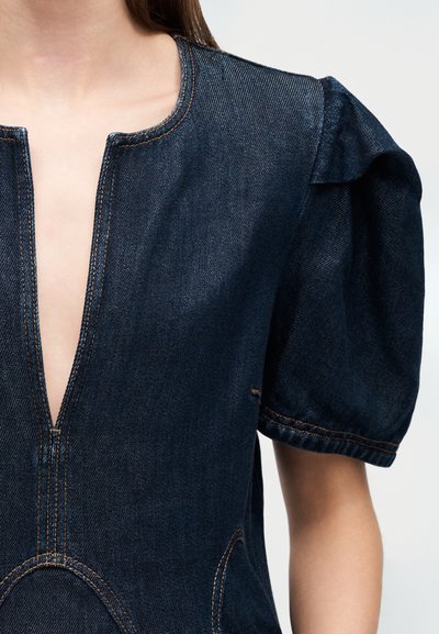 Τζιν τοπ με βαθύ ντεκολτέ σε σχήμα V, κοντά φουσκωτά μανίκια και ορατές ραφές. Το ύφασμα είναι σκούρο μπλε με διακριτική υφή και καμπύλες λεπτομέρειες στις ραφές.