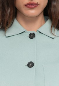 Lichte mintgroene wollen jas met een kraag, zwarte knopen en een voorzak. Gladde textuur met een klassiek ontwerp.