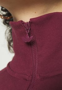 Sudadera burdeos con cremallera y cuello alto, textura acanalada, y un tirador de cremallera con un logo de tres rayas.