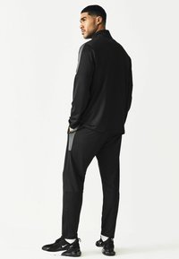 Musta urheilullinen track suit, jossa on pitkät hihat, vetoketjukaulus ja sivusaumapussit. Harmaat ja mustat kengät, joissa on teksturoitu yksityiskohta.
