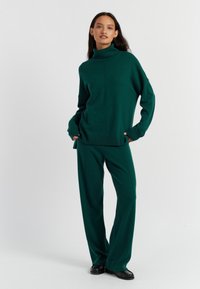 Maglione verde a collo alto in maglia abbinato a pantaloni larghi coordinati. Presenta polsini e orli a costine, con una texture liscia su tutta la superficie.