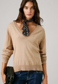 Bež pulover s pletenjem in V-izrezom, z rebrastim robom in manšetami, oblečen z črno-bež šahovnico, v kombinaciji z jeans hlačami in temno torbico.