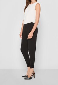 Biała bluzka bez rękawów z teksturowanym dekoltem, zestawiona z czarnymi zwężanymi spodniami z marszczeniami na kostkach, dopełniona czarnymi szpilkami.