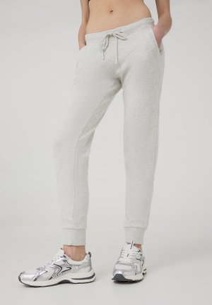 Pantaloni della tuta grigio chiaro con cintura regolabile, tasche laterali e polsini affusolati, abbinati a scarpe da ginnastica argento e bianche.