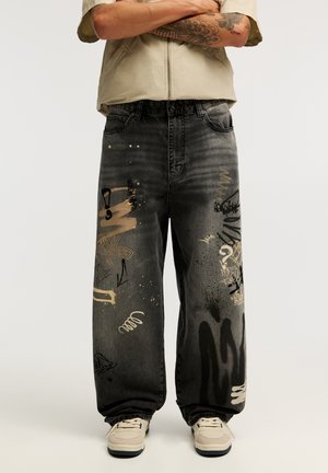 Personne portant un jean noir ample avec des éclaboussures de peinture abstraites et des motifs graffiti, des baskets claires et une chemise beige à manches courtes.