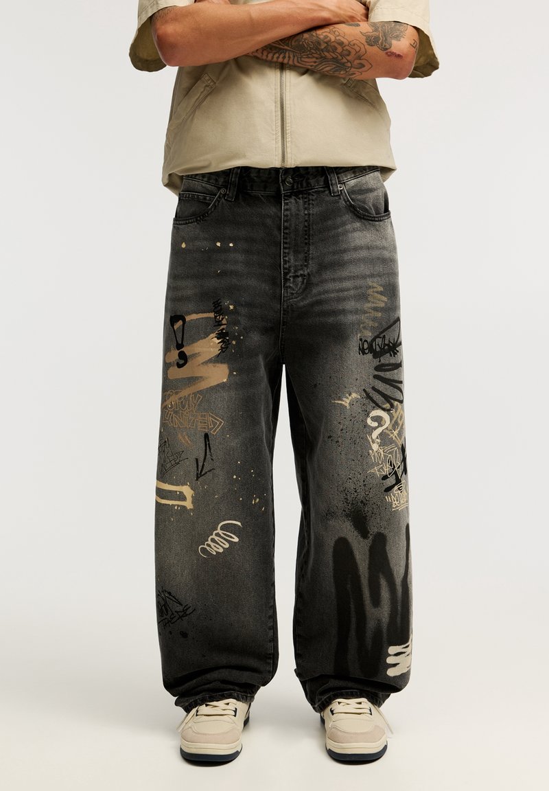 Personne portant un jean noir ample avec des éclaboussures de peinture abstraites et des motifs graffiti, des baskets claires et une chemise beige à manches courtes.