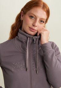Felpa con zip di colore viola tenue, dotata di un alto colletto, zip frontale e decorata con strass che formano la scritta "FREDDY". Texture morbida.