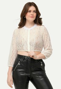 Top corto in pizzo con colletto bianco a ondine, maniche lunghe trasparenti e plissettature frontali. Abbinato a pantaloni alti in similpelle neri.