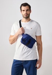 Marineblå nylon crossbody taske med justerbar rem, frontlynlås lomme og sorte hardware detaljer, båret af en person i en hvid skjorte.