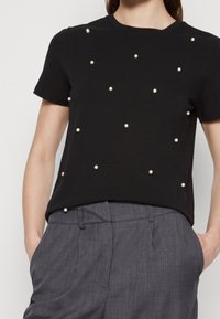 Camiseta negra de manga corta con adornos de perlas blancas dispersos, escote redondo y un corte relajado, combinada con pantalones grises.
