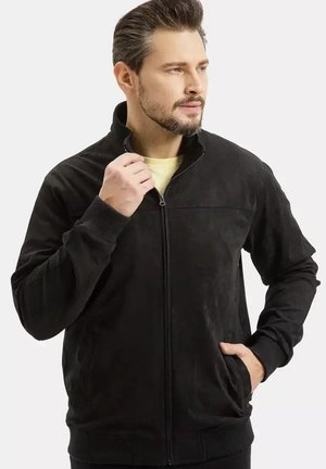 Mężczyzna z brodą, ubrany w czarną kurtkę na zamek, prawą ręką w kieszeni, lewą ręką poprawiający kołnierz, patrzący lekko w prawo.