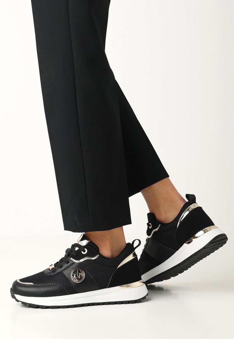 Personne portant des baskets noires avec semelles blanches et accents dorés, associée à un pantalon noir, debout sur un fond blanc uni.