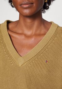 V-Ausschnitt Strickpullover in Hellbraun mit gerippter Textur und kleinem Logoakzent auf der Brust.