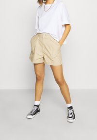 Žena oblečená v béžových šortkách, bílé oversized tričko, černých vysokých teniskách s bílými ponožkami, stojící na jednolitém světlem pozadí.