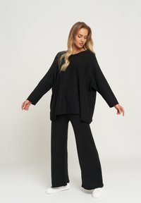 Czarny oversize sweter i pasujące szerokie spodnie z teksturowanej tkaniny, w połączeniu z białymi sneakersami. Neutralne tło.
