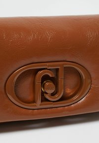 Sac à main en cuir synthétique marron avec un détail de quincaillerie logo brillant. Le sac a une texture lisse avec un motif de grain subtil.